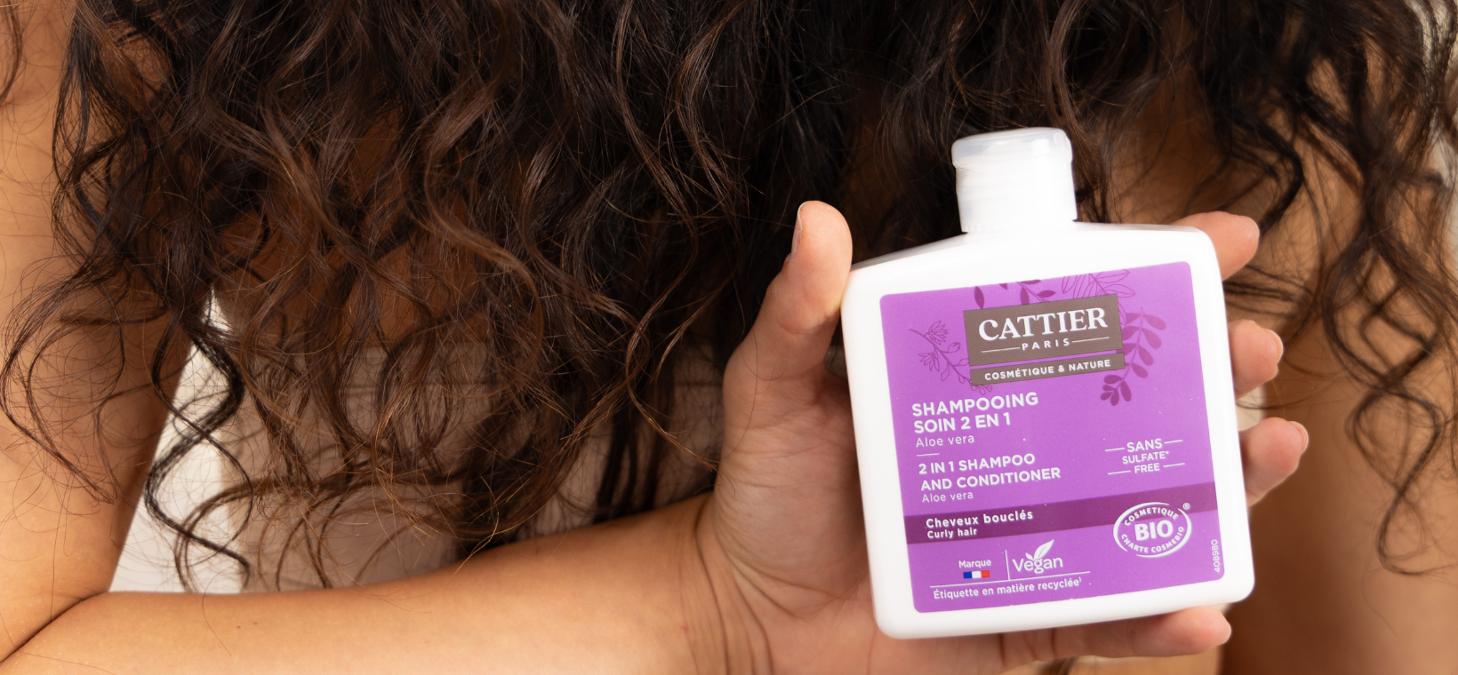 Visuel d'une femme aux cheveux bouclés tenant le shampoing soin cheveux bouclés bio Cattier